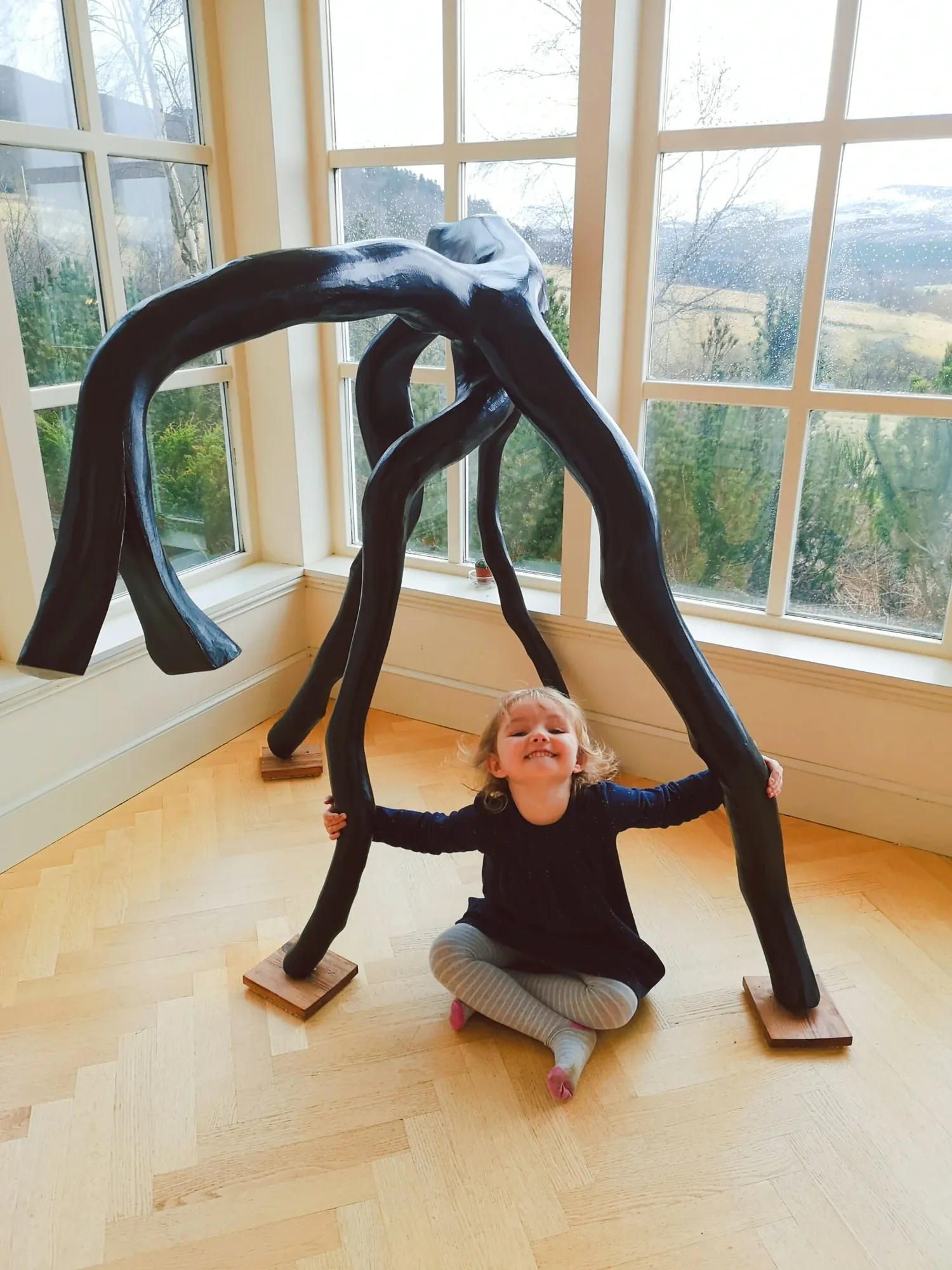 Horse, a large carved wood sculpture by Bruce Thomson, a black form arching over on splayed legs, with Bruce's granddaughter sitting beneath it for scale