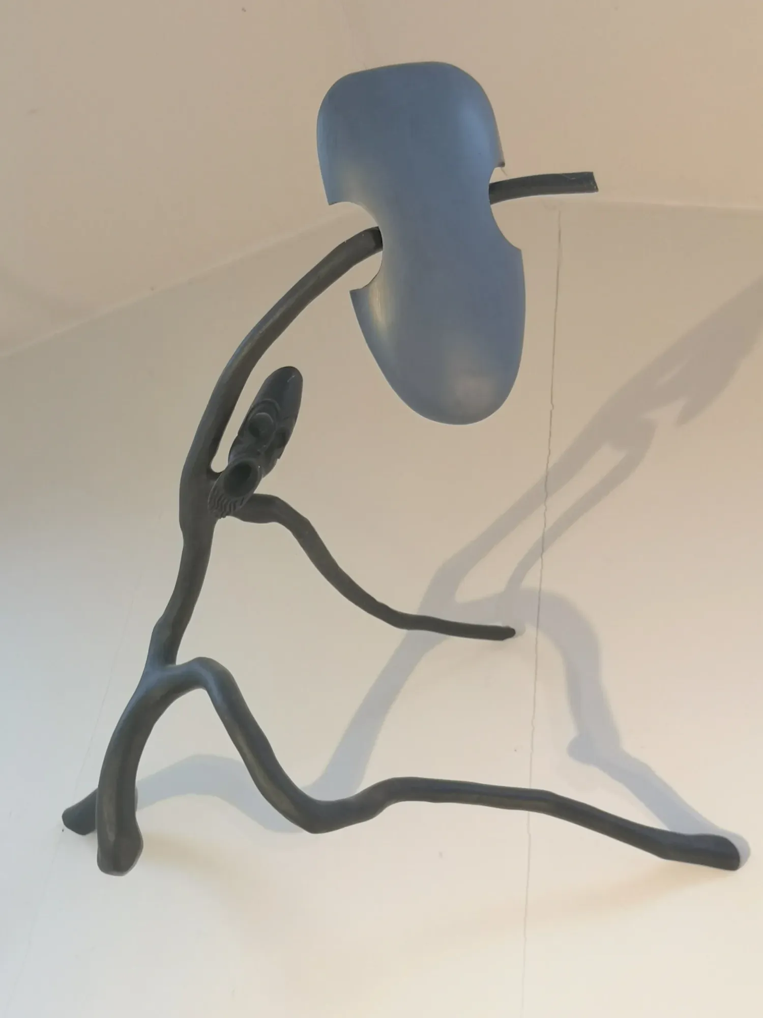 Warrior, a carved wood sculpture by Bruce Thomson, a lean figure crouched behind a large pale shield with a spear